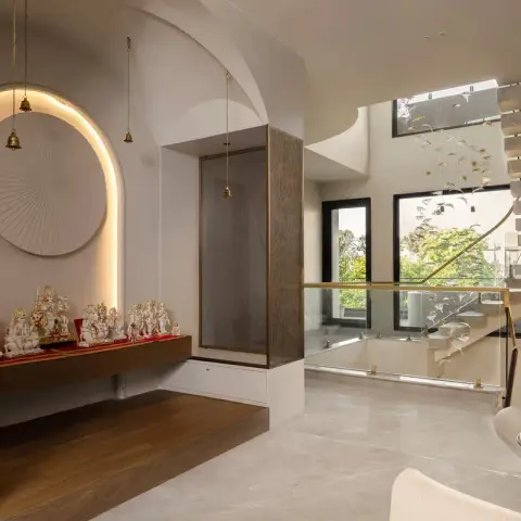 Arched pooja niche lit by a concealed halo, with white marble idols on a floating teak ledge, brass bells hung on chains and